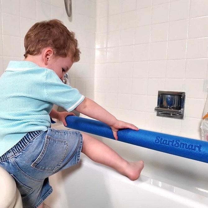 A young child in a light blue shirt and denim shorts tries to climb on the edge of a white bathtub, holding onto a blue StartSmart pool noodle in a tiled bathroom. He can't get over the tub with the safety bar in place. 