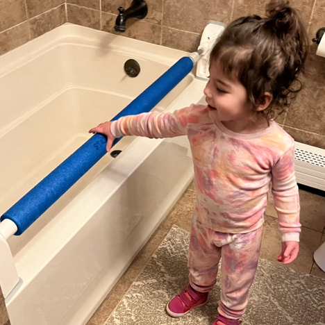 A toddler in a tie-dye pajama set standing besides a white bathtub, resting her hand on the blue foam covered StartSmart Safety Bar that is mounted to the tub's edge. 