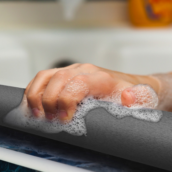 A close-up of a hand gripping the gray safety bar, covered in white foam, with a blurred sink and orange bottle in the background.